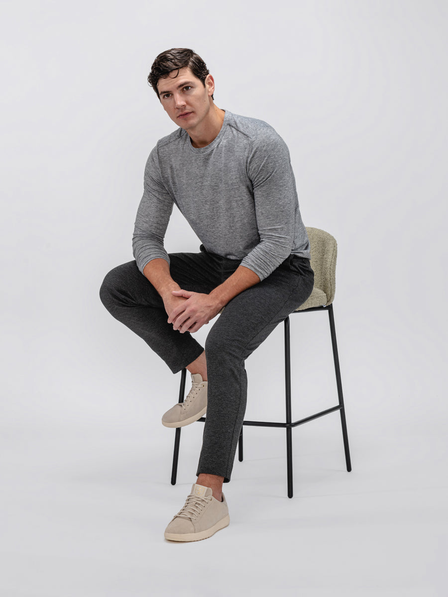 model wearing men's pale grey heather composite merino long sleeve tee and charcoal fusion pant sitting in chair and leaning forward with sleeves rolled up