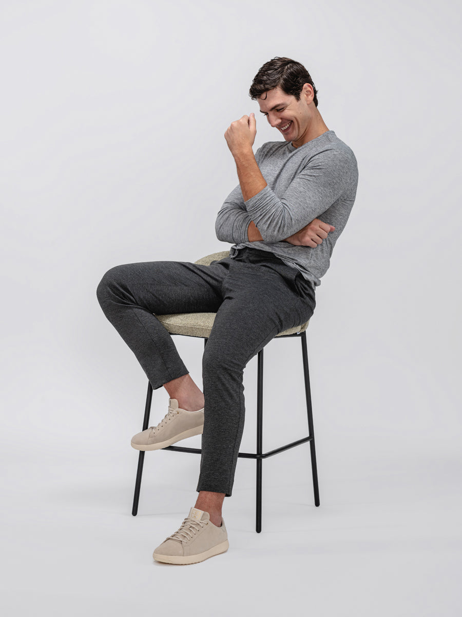 model wearing men's pale grey heather composite merino long sleeve tee and charcoal fusion pant sitting in chair and leaning back with arms crossed and sleeves rolled up